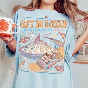 May include: Light blue t-shirt with the text "GET IN LOSER WE'RE GOING TO SPACE" and a graphic of a space-themed amusement park ride with cartoon characters. The design includes planets, stars, and flying saucers.