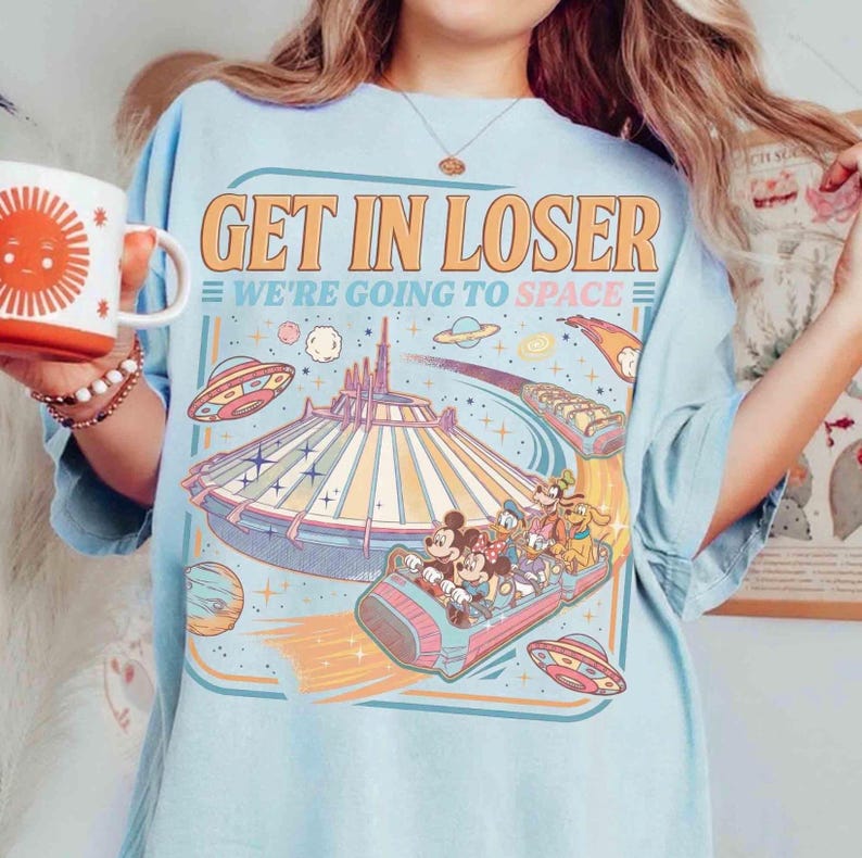May include: Light blue t-shirt with the text "GET IN LOSER WE'RE GOING TO SPACE" and a graphic of a space-themed amusement park ride with cartoon characters. The design includes planets, stars, and flying saucers.