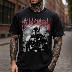 May include: Black t-shirt featuring a graphic of The Mandalorian with a helmet and weapon, set against a backdrop of lightning. The text "THE MANDALORIAN" is displayed in red, stylized lettering above the image.