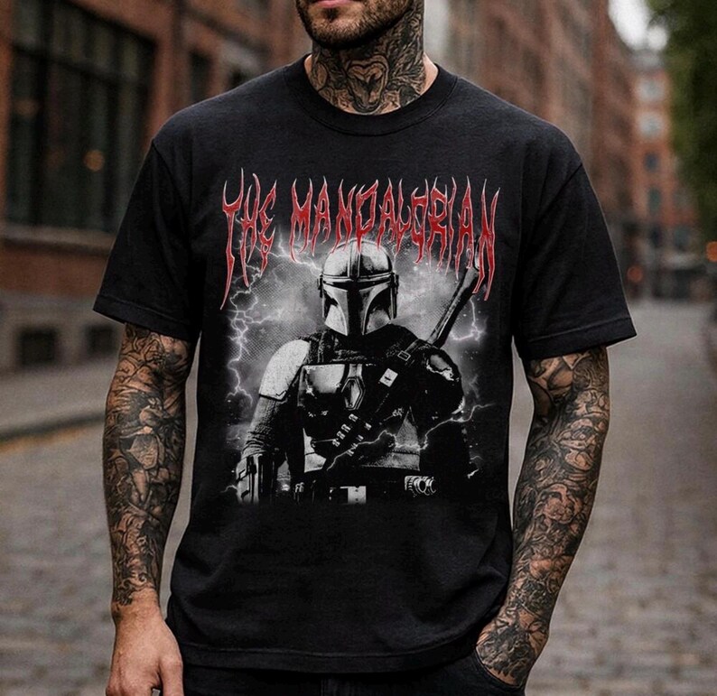 May include: Black t-shirt featuring a graphic of The Mandalorian with a helmet and weapon, set against a backdrop of lightning. The text "THE MANDALORIAN" is displayed in red, stylized lettering above the image.
