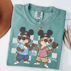 Peut inclure: T-shirt Comfort Colors bleu sarcelle avec des personnages de dessins anim&eacute;s Chip et Dale portant des lunettes de soleil et des chapeaux Mickey Mouse, tenant des glaces. Le t-shirt a un fond &agrave; carreaux et les mots "Chip" et "Dale". Un collier argent&eacute; est drap&eacute; sur le t-shirt.