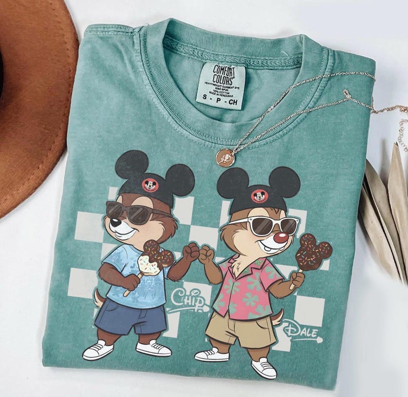 Peut inclure: T-shirt Comfort Colors bleu sarcelle avec des personnages de dessins anim&eacute;s Chip et Dale portant des lunettes de soleil et des chapeaux Mickey Mouse, tenant des glaces. Le t-shirt a un fond &agrave; carreaux et les mots "Chip" et "Dale". Un collier argent&eacute; est drap&eacute; sur le t-shirt.