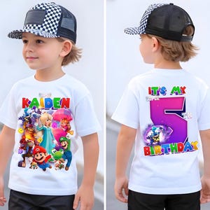 May include: A white t-shirt with a vibrant graphic of video game characters and the name "KAIDEN" in red, blue, and green. The back of the shirt says "IT'S MY SUPER 5TH BIRTHDAY" with a large purple number 5. The child is wearing a black and white checkered cap.