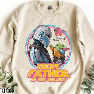 May include: A natural-colored sweatshirt featuring a graphic of The Mandalorian and Grogu, with the text "BEST FATHER IN THE GALAXY" in yellow and pink. The design is set within a pink and blue circle, with a sunset graphic.