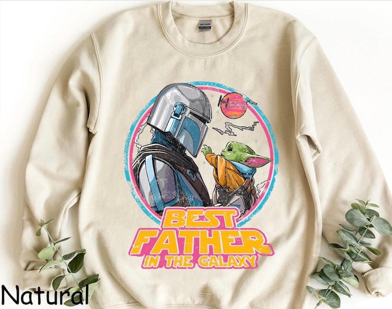 May include: A natural-colored sweatshirt featuring a graphic of The Mandalorian and Grogu, with the text "BEST FATHER IN THE GALAXY" in yellow and pink. The design is set within a pink and blue circle, with a sunset graphic.