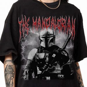 May include: Black t-shirt featuring a graphic of The Mandalorian with a helmet and weapon, set against a stormy background. The text "THE MANDALORIAN" is printed in red, gothic-style lettering across the top.