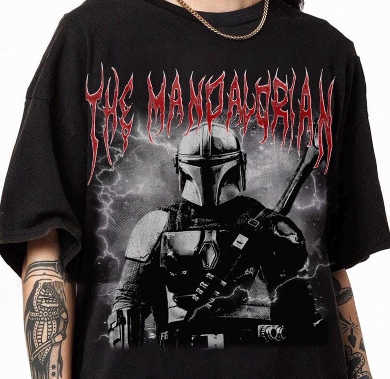 May include: Black t-shirt featuring a graphic of The Mandalorian with a helmet and weapon, set against a stormy background. The text "THE MANDALORIAN" is printed in red, gothic-style lettering across the top.