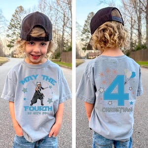 May include: A light blue t-shirt featuring the text "MAY THE FOURTH BE WITH YOU" and a character graphic with lightsabers. The back of the shirt displays a large number 4, fireworks, balloons, stars, and the name "CHRISTIAN".