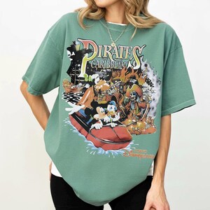 May include: Green t-shirt with a graphic from the Pirates of the Caribbean ride at Disneyland. The design includes Mickey Mouse, Donald Duck, and Goofy in a boat, with pirate imagery and the ride's logo. The shirt has a relaxed fit.