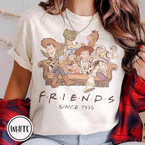 May include: White t-shirt featuring a graphic of Toy Story characters on a brown couch. The word "FRIENDS" is printed in black, with "SINCE 1995" below. The shirt has short sleeves and a crew neck.