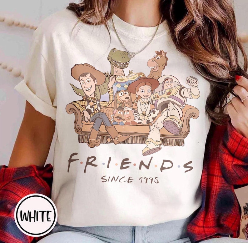 May include: White t-shirt featuring a graphic of Toy Story characters on a brown couch. The word "FRIENDS" is printed in black, with "SINCE 1995" below. The shirt has short sleeves and a crew neck.