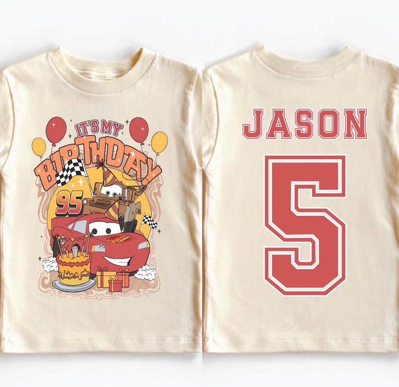 May include: A cream-colored t-shirt featuring a birthday design with the text "It's My Birthday" and the number "95". The design includes cartoon cars, balloons, and a birthday cake. The back of the shirt displays the name "JASON" and the number "5" in red.