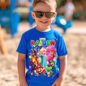 May include: A young boy wearing a blue t-shirt with the text "SUPER KAIDEN" in red and white. The shirt features a colourful graphic of video game characters. The boy is wearing sunglasses and smiling.