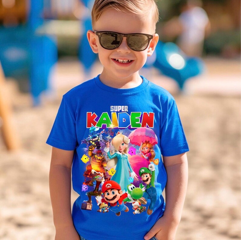 May include: A young boy wearing a blue t-shirt with the text "SUPER KAIDEN" in red and white. The shirt features a colourful graphic of video game characters. The boy is wearing sunglasses and smiling.