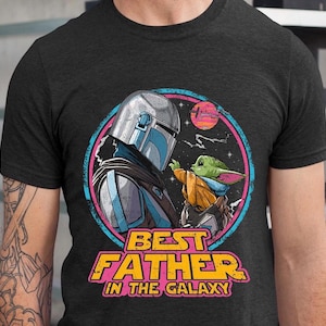 May include: Dark gray t-shirt featuring a colorful graphic of a Mandalorian character and Grogu, with the text "BEST FATHER IN THE GALAXY". The design includes a space scene with a sunset and stars.