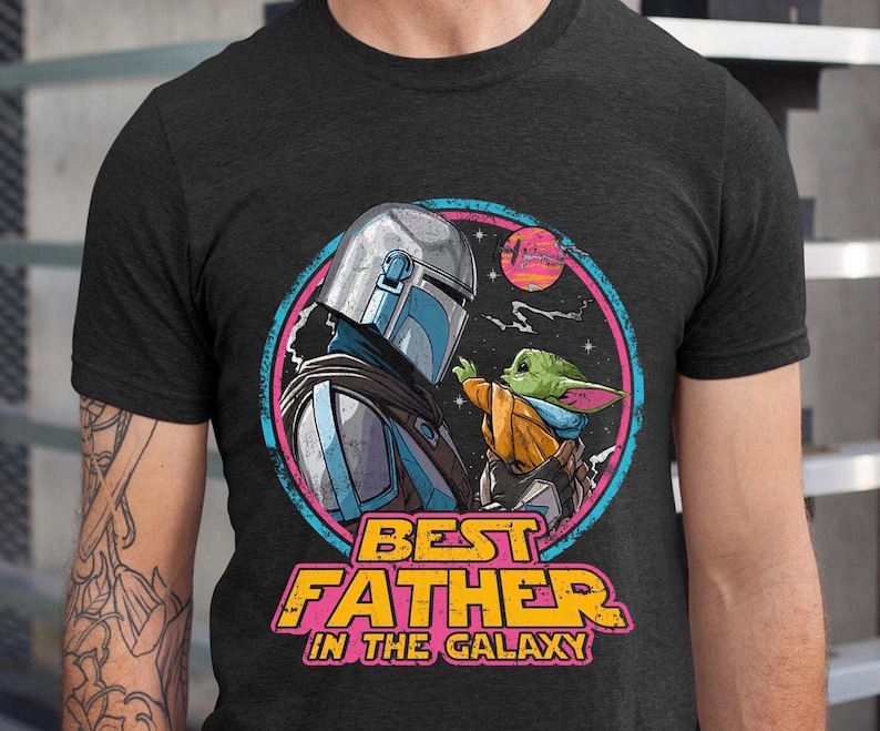 May include: Dark gray t-shirt featuring a colorful graphic of a Mandalorian character and Grogu, with the text "BEST FATHER IN THE GALAXY". The design includes a space scene with a sunset and stars.