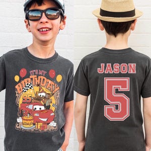 May include: Charcoal gray t-shirt with a colorful graphic featuring a red race car, birthday cake, and the text "IT'S MY BIRTHDAY". The back of the shirt displays the name "JASON" and the number "5" in red.