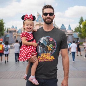 May include: A man wearing a dark gray t-shirt with a graphic of The Mandalorian and Grogu, with the text "BEST FATHER IN THE GALAXY". He is holding a young child wearing a red polka dot dress and Minnie Mouse ears.