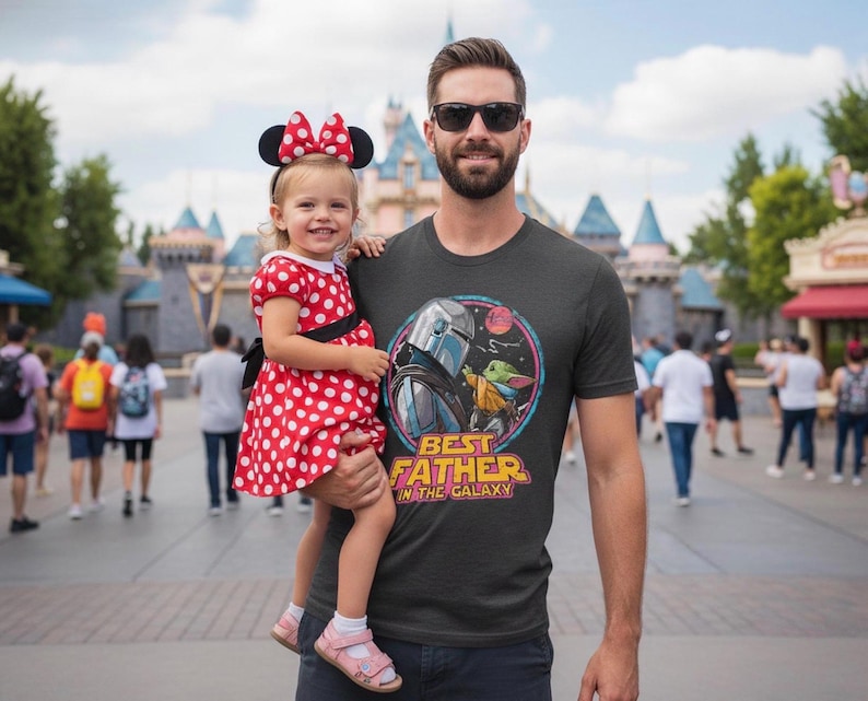 May include: A man wearing a dark gray t-shirt with a graphic of The Mandalorian and Grogu, with the text "BEST FATHER IN THE GALAXY". He is holding a young child wearing a red polka dot dress and Minnie Mouse ears.