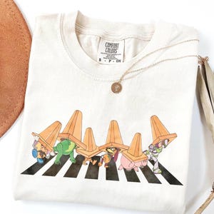 May include: Cream-coloured t-shirt with a graphic of cartoon characters crossing a zebra crossing, each wearing an orange traffic cone. The shirt has a Comfort Colors tag and a gold necklace.