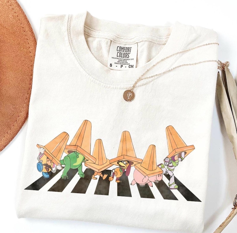 May include: Cream-coloured t-shirt with a graphic of cartoon characters crossing a zebra crossing, each wearing an orange traffic cone. The shirt has a Comfort Colors tag and a gold necklace.