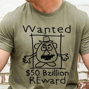K&ouml;nnte beinhalten: Olivgr&uuml;nes T-Shirt mit einer schwarzen Cartoon-Illustration eines kartoffelartigen Charakters in einem Fahndungsrahmen. Der Text "Wanted" steht &uuml;ber der Figur und "$50 Bzillion REWARD" darunter.