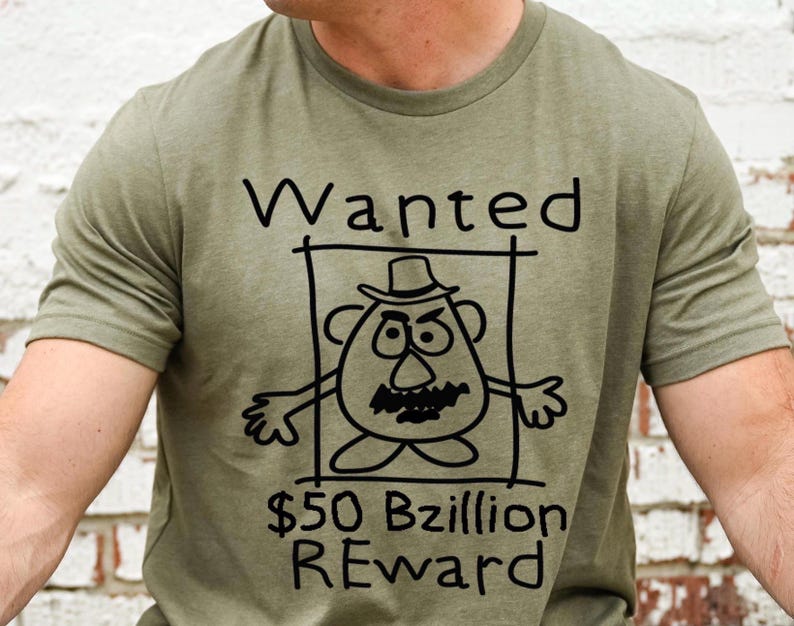 K&ouml;nnte beinhalten: Olivgr&uuml;nes T-Shirt mit einer schwarzen Cartoon-Illustration eines kartoffelartigen Charakters in einem Fahndungsrahmen. Der Text "Wanted" steht &uuml;ber der Figur und "$50 Bzillion REWARD" darunter.