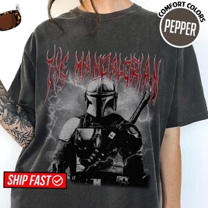 May include: Dark gray t-shirt featuring a graphic of The Mandalorian in black and white, with red text above. The shirt has a vintage, distressed look. The text "COMFORT COLORS PEPPER" is in a circle on the right side of the shirt.
