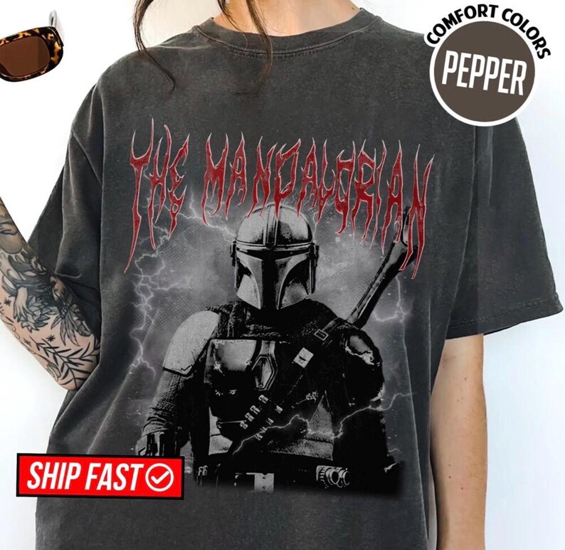 May include: Dark gray t-shirt featuring a graphic of The Mandalorian in black and white, with red text above. The shirt has a vintage, distressed look. The text "COMFORT COLORS PEPPER" is in a circle on the right side of the shirt.