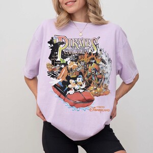 May include: A lilac t-shirt with a vibrant graphic of the Pirates of the Caribbean ride at Tokyo Disneyland. The design includes Mickey Mouse, Donald Duck, and a pirate ship. The text "Pirates of the Caribbean" and "Tokyo Disneyland" are also visible.