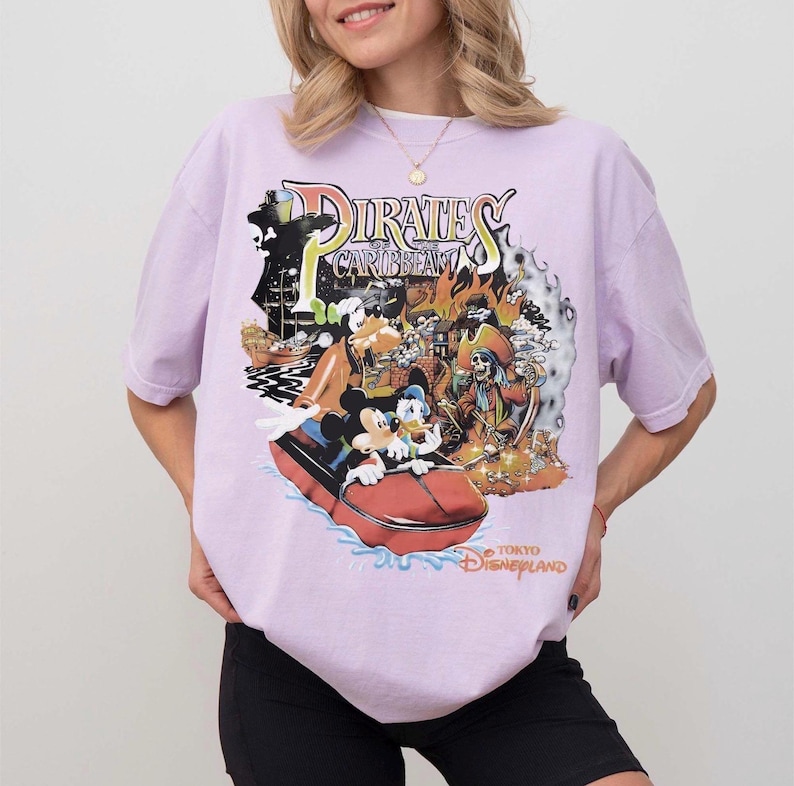 May include: A lilac t-shirt with a vibrant graphic of the Pirates of the Caribbean ride at Tokyo Disneyland. The design includes Mickey Mouse, Donald Duck, and a pirate ship. The text "Pirates of the Caribbean" and "Tokyo Disneyland" are also visible.