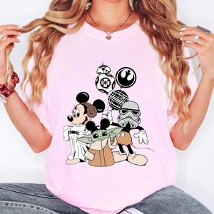 May include: A light pink t-shirt featuring a graphic of Mickey Mouse dressed as Princess Leia, Baby Yoda, and a Stormtrooper, with Star Wars themed balloons. The shirt is a crew neck style.