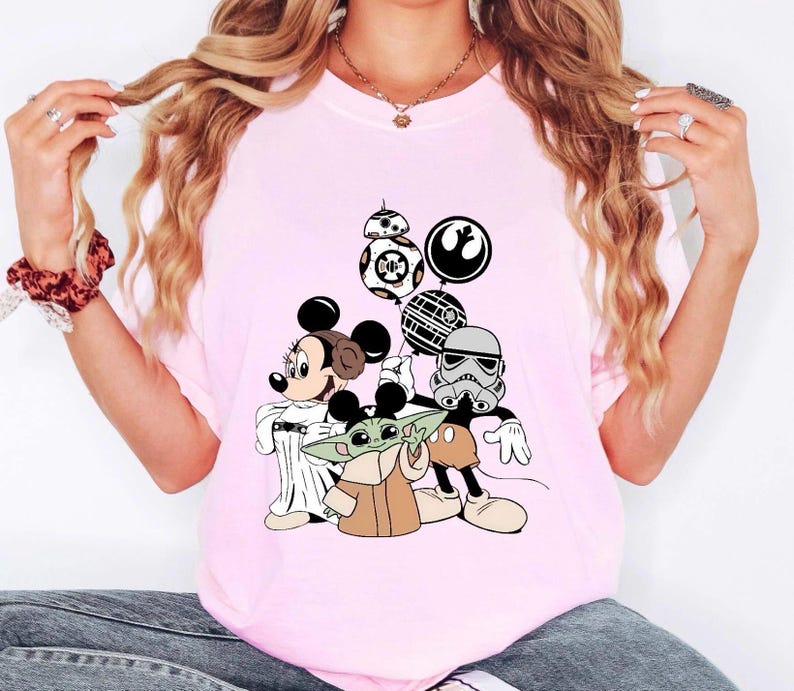 May include: A light pink t-shirt featuring a graphic of Mickey Mouse dressed as Princess Leia, Baby Yoda, and a Stormtrooper, with Star Wars themed balloons. The shirt is a crew neck style.