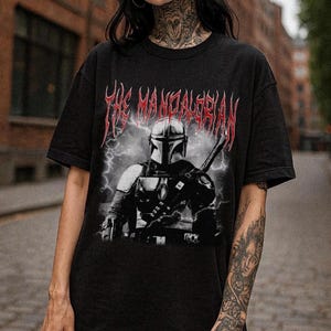 May include: Black t-shirt featuring a graphic of The Mandalorian with a helmet and weapon, set against a backdrop of lightning. The text "The Mandalorian" is displayed in red, stylized lettering above the image. The shirt is a dark color.