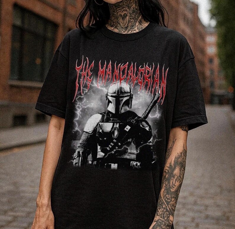 May include: Black t-shirt featuring a graphic of The Mandalorian with a helmet and weapon, set against a backdrop of lightning. The text "The Mandalorian" is displayed in red, stylized lettering above the image. The shirt is a dark color.