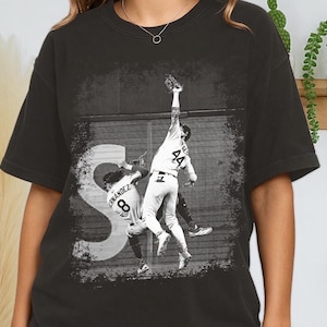 以下が含まれることがあります： 野球選手のアクションショットが描かれた、グレーのTシャツ。背番号44の選手がボールをキャッチしようとジャンプしています。背番号8の選手も写っています。選手の背後には大きな白い「S」があります。