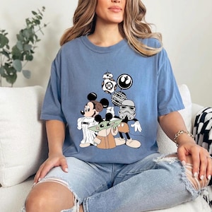 May include: A light blue t-shirt featuring a graphic of Mickey Mouse, Baby Yoda, and a Stormtrooper. The design includes Star Wars and Disney characters. The shirt is a comfortable, casual style.