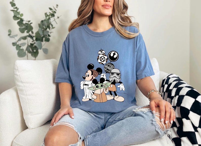 May include: A light blue t-shirt featuring a graphic of Mickey Mouse, Baby Yoda, and a Stormtrooper. The design includes Star Wars and Disney characters. The shirt is a comfortable, casual style.