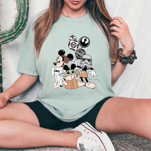 May include: Light green t-shirt featuring a graphic of Mickey Mouse, Baby Yoda, and Star Wars characters. The design includes BB-8, a Stormtrooper, and other iconic elements from the Star Wars franchise. The shirt is a comfortable, casual style.