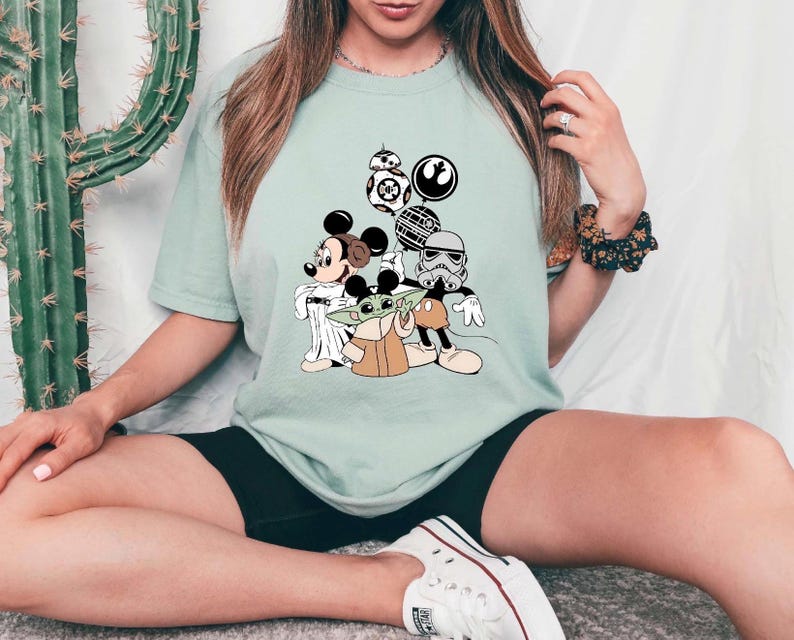 May include: Light green t-shirt featuring a graphic of Mickey Mouse, Baby Yoda, and Star Wars characters. The design includes BB-8, a Stormtrooper, and other iconic elements from the Star Wars franchise. The shirt is a comfortable, casual style.