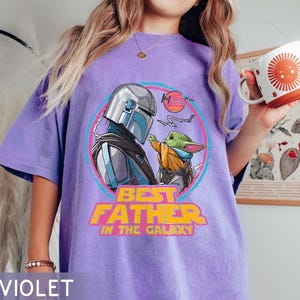 May include: Violet oversized t-shirt featuring a graphic of The Mandalorian and Grogu, with the text "Best Father in the Galaxy." The design is set within a pink and blue circle. A white mug with an orange sun design is held in the hand.