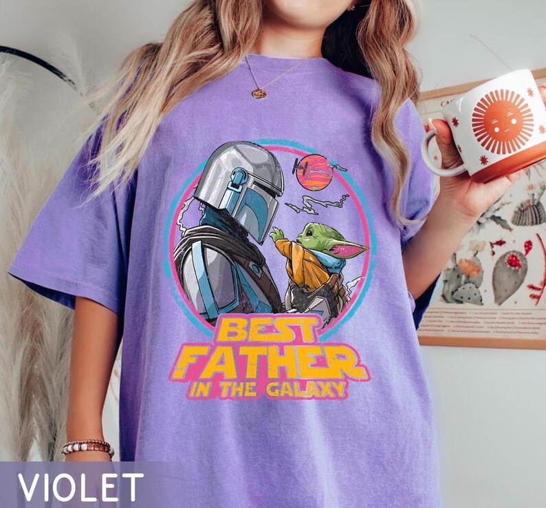 May include: Violet oversized t-shirt featuring a graphic of The Mandalorian and Grogu, with the text "Best Father in the Galaxy." The design is set within a pink and blue circle. A white mug with an orange sun design is held in the hand.