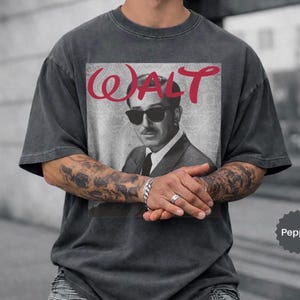 May include: Dark gray t-shirt featuring a black and white image of a man wearing sunglasses, with the name "WALT" in red script above the image. The shirt has short sleeves and a relaxed fit.