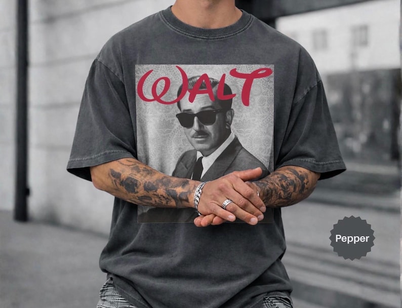 May include: Dark gray t-shirt featuring a black and white image of a man wearing sunglasses, with the name "WALT" in red script above the image. The shirt has short sleeves and a relaxed fit.