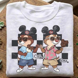 Peut inclure: Sweat-shirt blanc &agrave; col rond avec une illustration de dessin anim&eacute; de Tic et Tac portant des lunettes de soleil et des chapeaux de Mickey Mouse. Ils tiennent des glaces enrob&eacute;es de chocolat. Les mots "Chip" et "Dale" sont imprim&eacute;s.