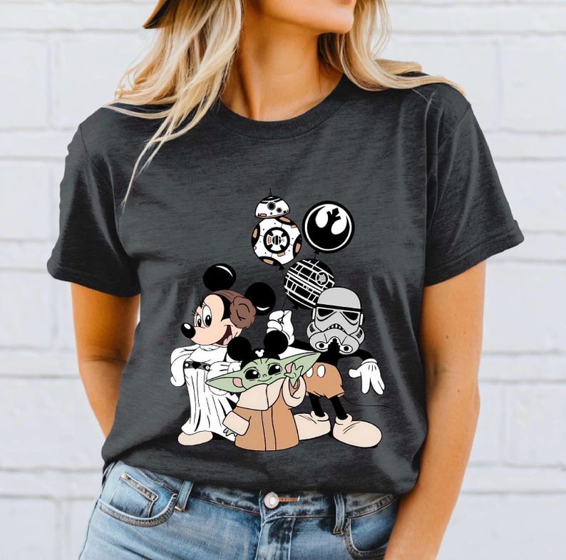 May include: Dark gray t-shirt featuring a graphic of Mickey Mouse, Minnie Mouse, Grogu, and Star Wars characters. The design includes BB-8, a Stormtrooper, and the Rebel Alliance symbol. The characters are in a cartoon style.