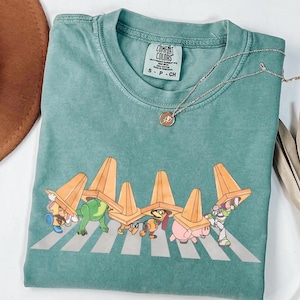 May include: A teal Comfort Colors t-shirt with a graphic of Toy Story characters crossing a zebra crossing, wearing orange traffic cones. A silver necklace with a small pendant is on the shirt.