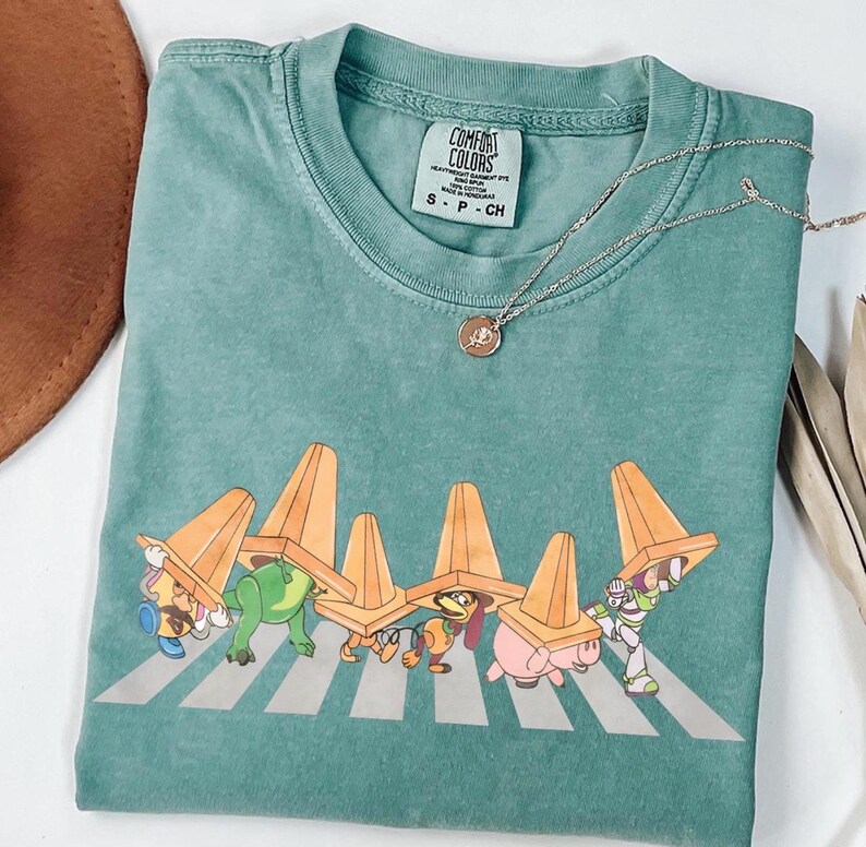 May include: A teal Comfort Colors t-shirt with a graphic of Toy Story characters crossing a zebra crossing, wearing orange traffic cones. A silver necklace with a small pendant is on the shirt.