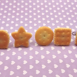 Puede incluir: Un conjunto de cinco pendientes de botón con forma de galletas doradas en miniatura. Las formas incluyen un corazón, una estrella de cinco puntas, un círculo, un cuadrado y un pequeño rectángulo, cada uno con pequeñas hendiduras y postes metálicos.