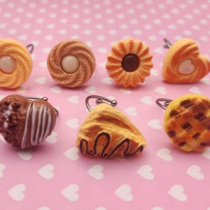 May include: A collection of seven miniature cookie-shaped rings featuring various designs including swirled biscuits with centers, a flower-shaped cookie, heart-shaped treats, a chocolate-drizzled wedge, and a lattice-topped round cookie.